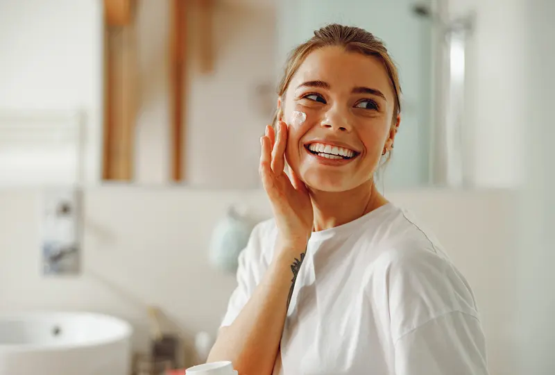 A woman in a white shirt smiles while applying cream to her face in a bathroom setting.