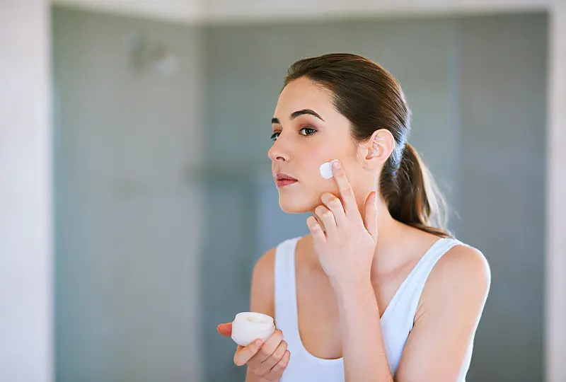 Woman applying face cream while looking in the mirror, wearing a white tank top, with blurred bathroom background.