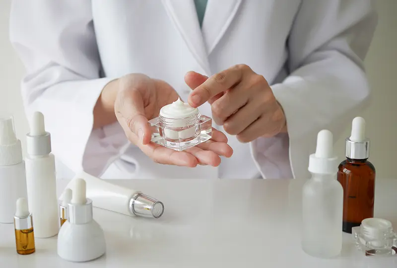 Person in a white coat holding a jar of cream, surrounded by various cosmetic bottles and containers on a white table.