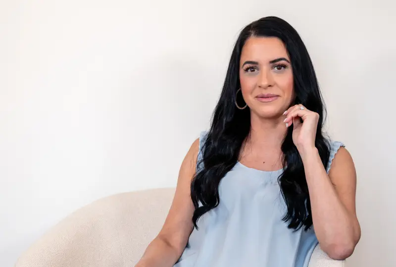 Woman with long dark hair sitting in a light-colored chair, wearing a light blue sleeveless top, looking at the camera.