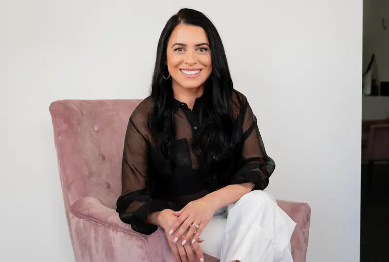 A woman with long black hair sits on a pink chair, wearing a sheer black top and white pants. She is smiling with hands resting on her lap.
