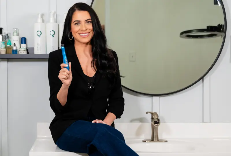 Person sitting on a bathroom counter holding a blue bottle, with various toiletries around and a round mirror in the background.