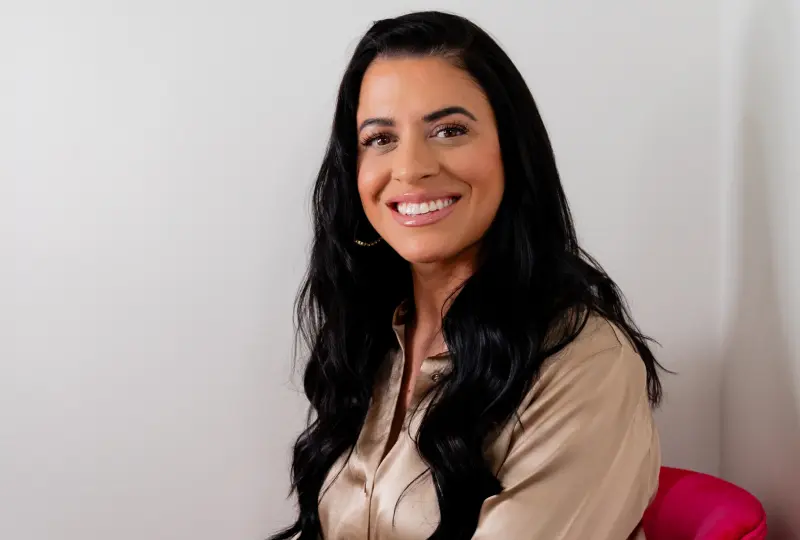 A woman with long black hair, wearing a beige blouse, smiling and sitting against a white background.