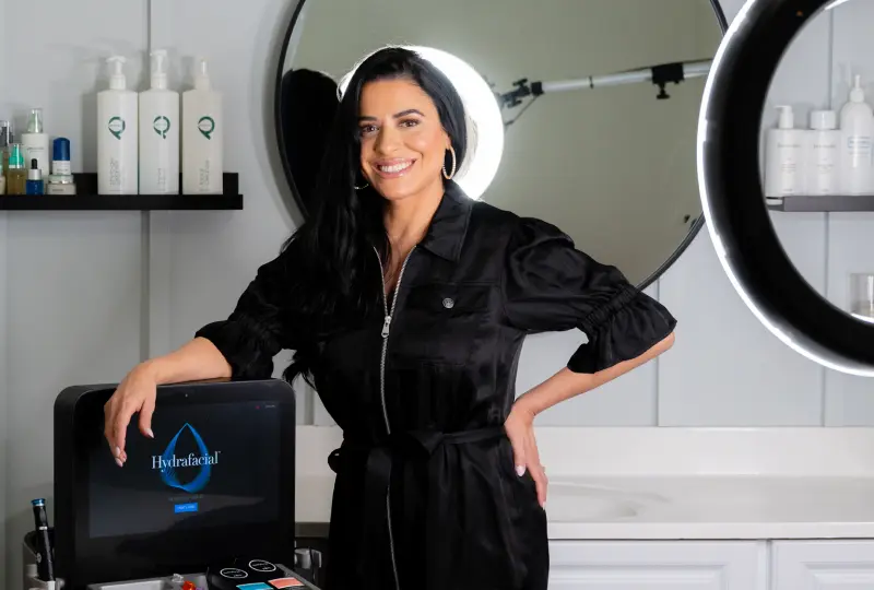 Person in a black outfit stands beside skincare equipment, smiling. Shelves with beauty products are visible in the background.