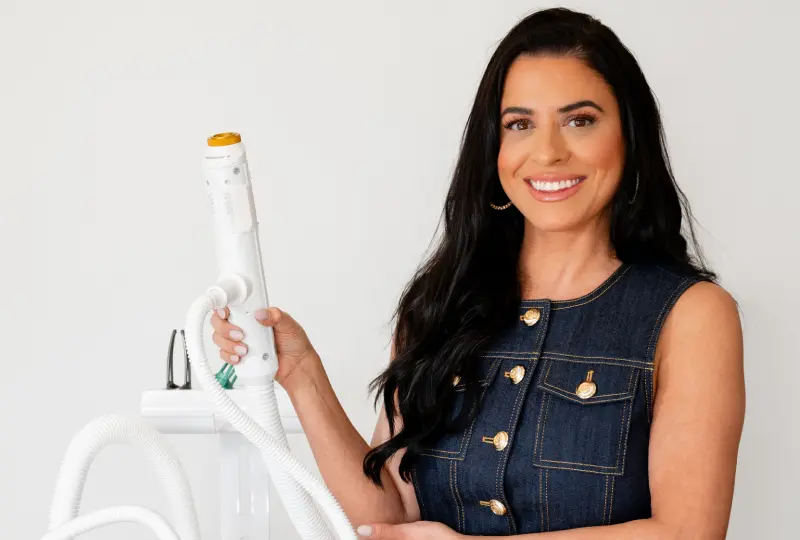 A person with long dark hair smiles while holding a white medical device with multiple cords against a plain background.
