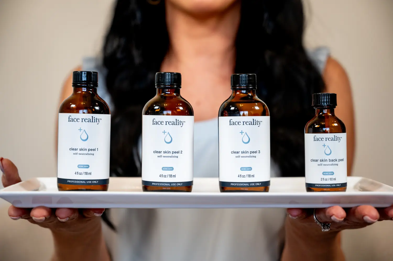 GoDaddy__LAW1537 Person holding a white tray with four bottles labeled "face reality" clear skin peels, numbered 1 to 3, and a back peel.
