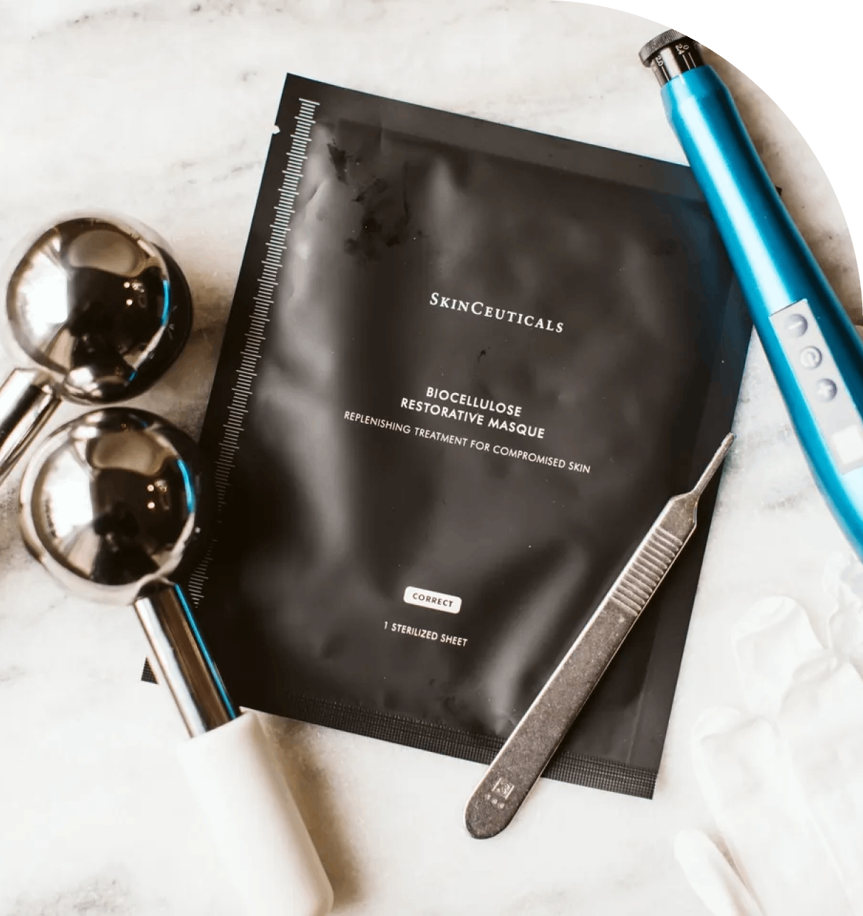 gghGroup 1321317369 A black skincare mask packet alongside a blue device, two metallic rollers, a pair of gloves, and a tweezer on a marble surface.