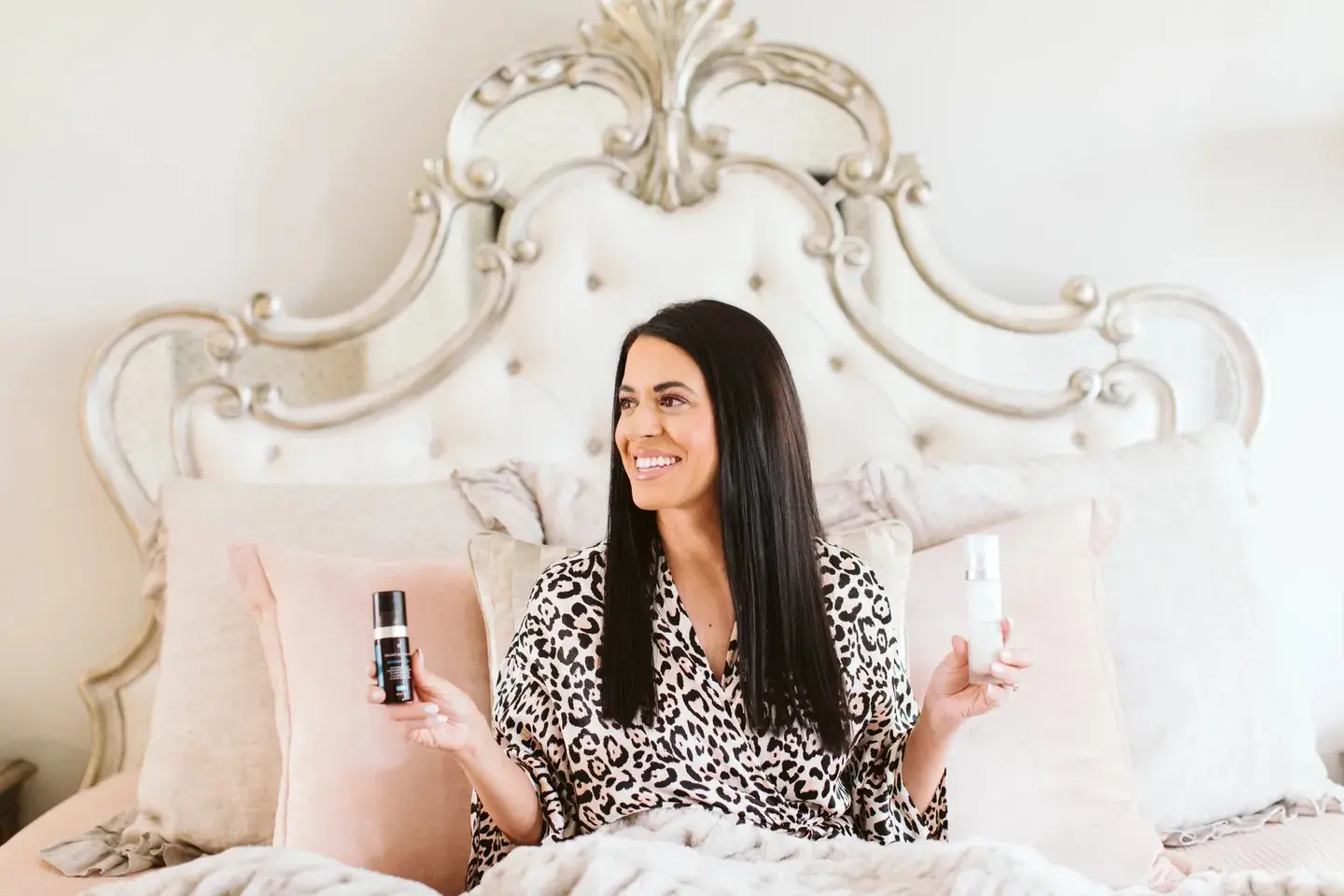 hero-img3 A woman in a leopard print robe sits on a bed holding two bottles, smiling. The bed has a decorative headboard and multiple pillows.