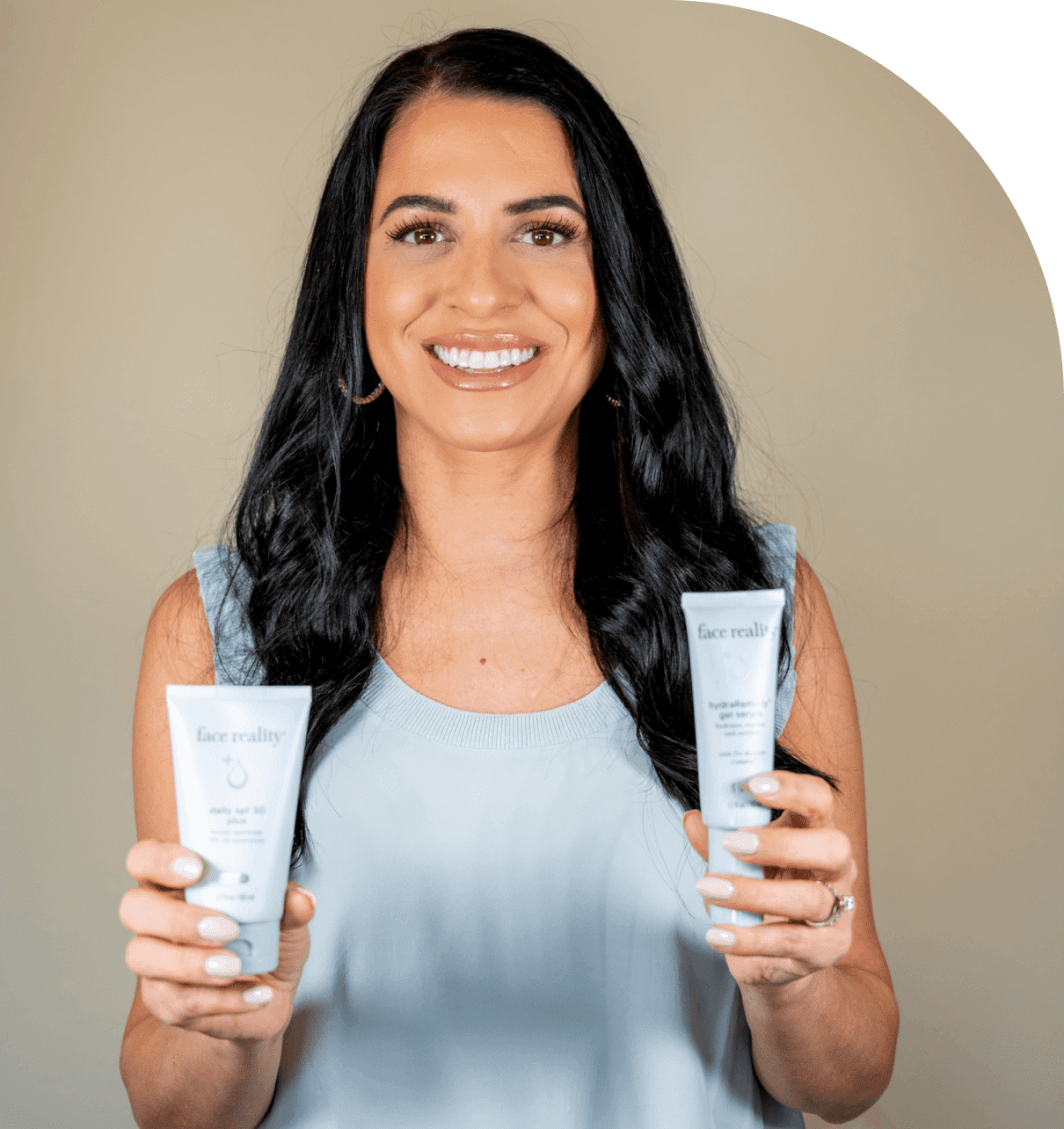 Group 1321317374 A woman with long dark hair, wearing a light blue sleeveless top, smiles while holding two skincare product tubes labeled "face reality.